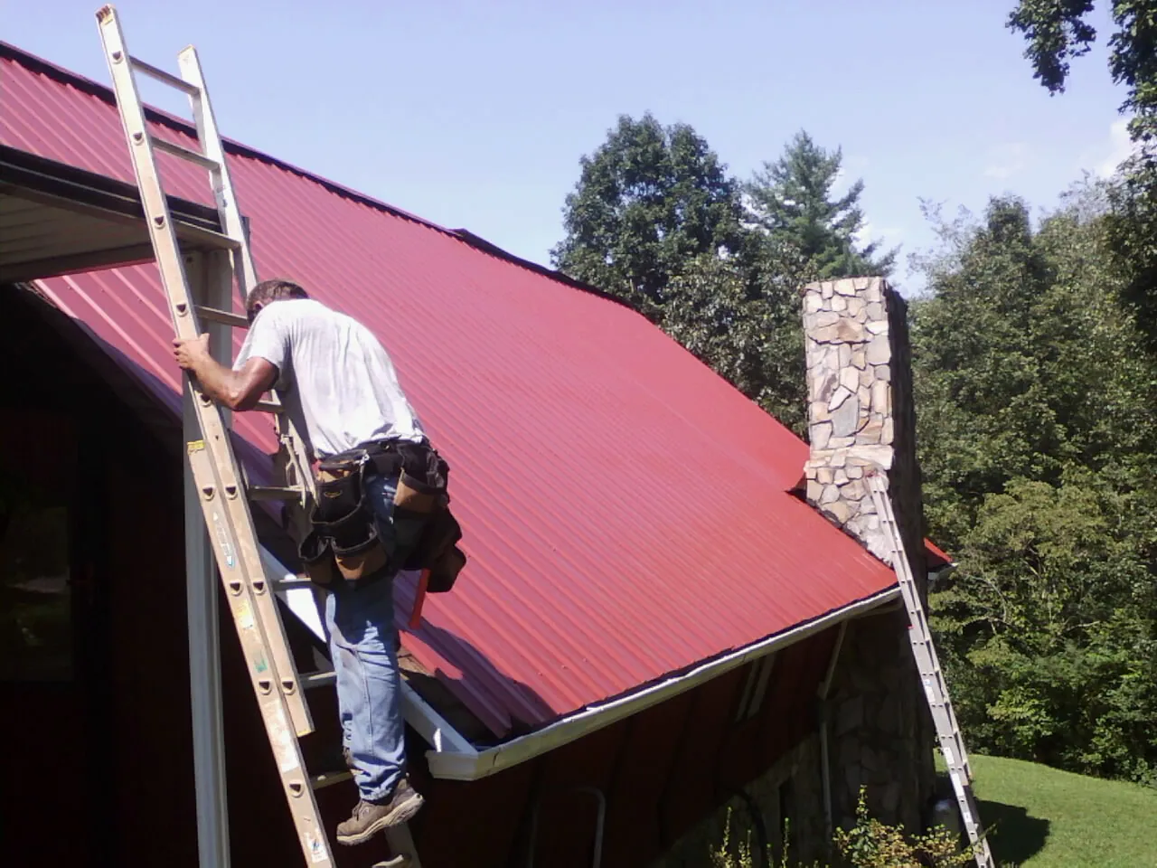 Roofer on a ladder beside a red metal roof and stone chimney — Soffit & Fascia Repair by Roofing Brothers in Newbury